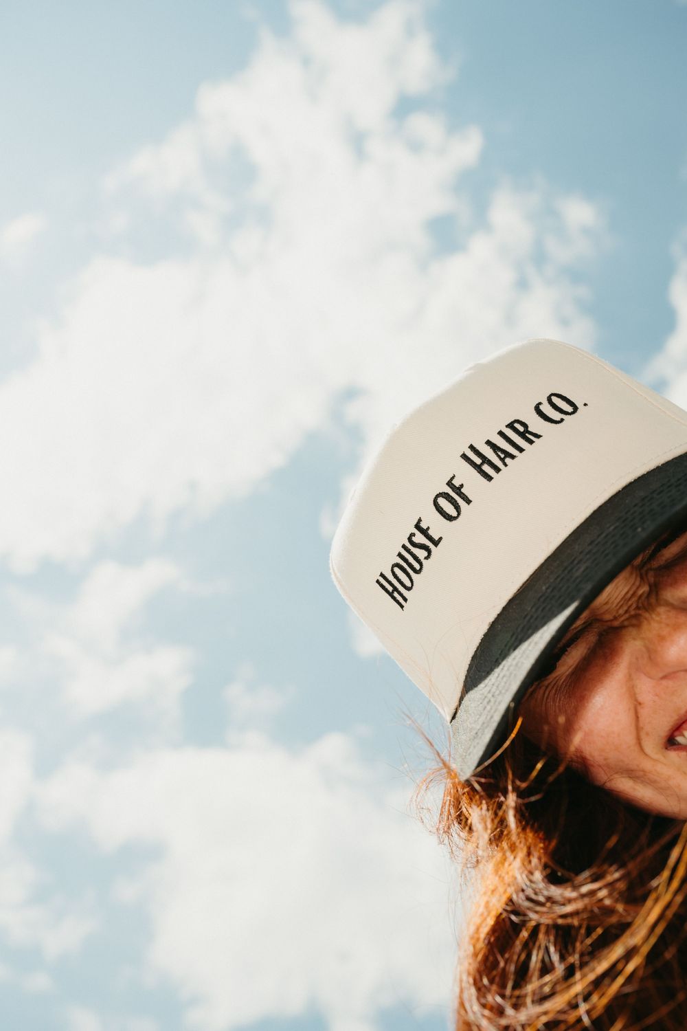 Person wearing a white baseball cap gazes up at blue sky with white fluffy clouds on a sunny day.