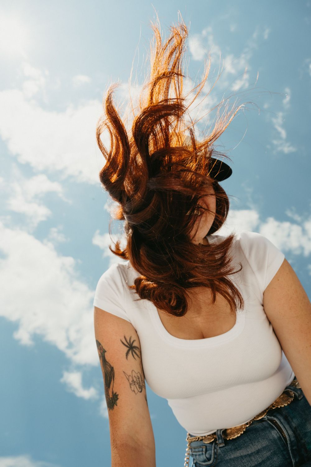 Long brown hair blowing in the wind against blue sky, wearing white tank top and denim with visible arm tattoos.