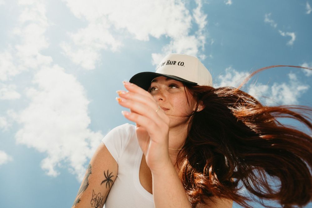 A person in a white tank top and baseball cap poses against a blue sky with wispy clouds as their long hair flows in the wind.