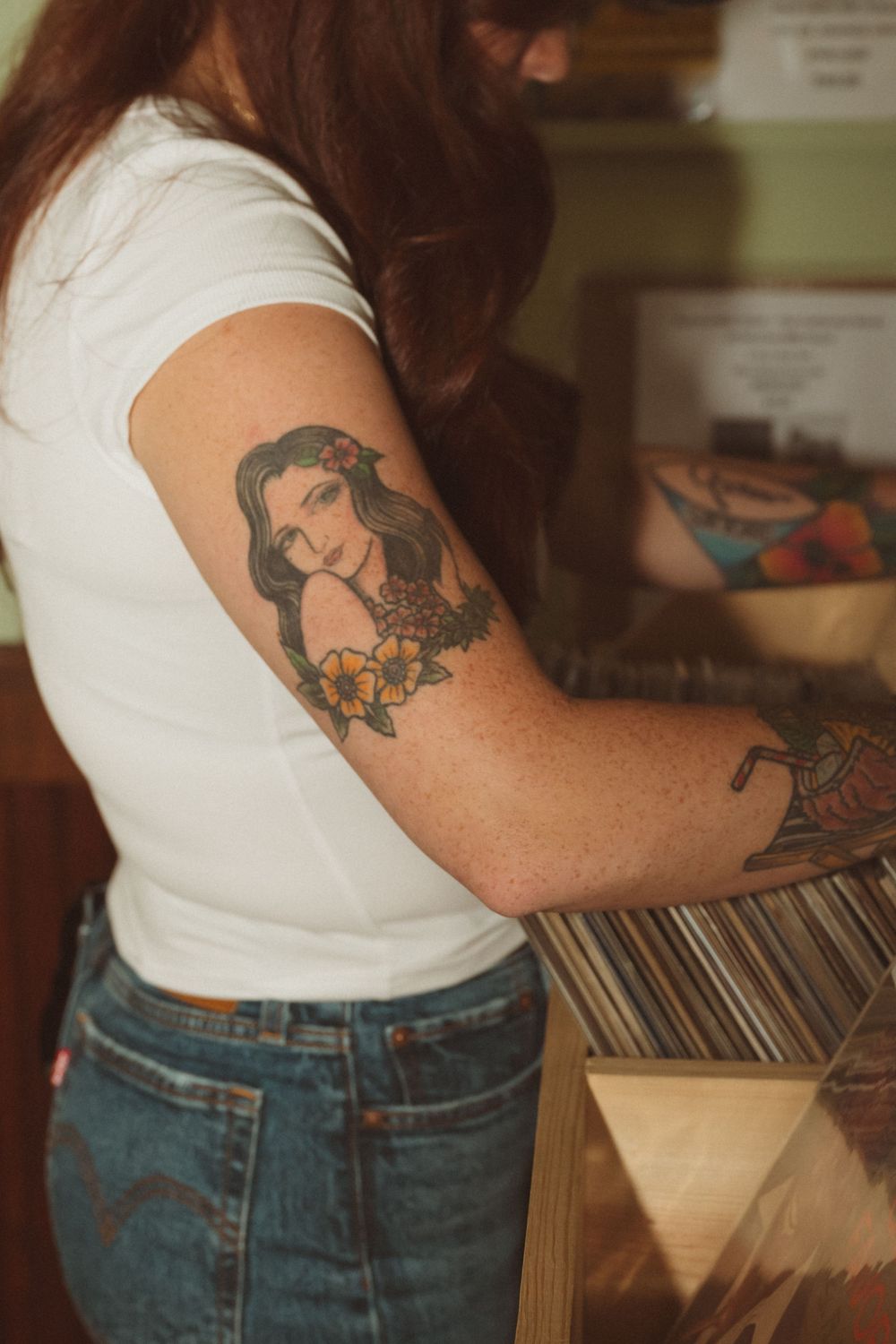 Person browsing through vinyl records in a record store wearing jeans and a white t-shirt with visible tattoos.