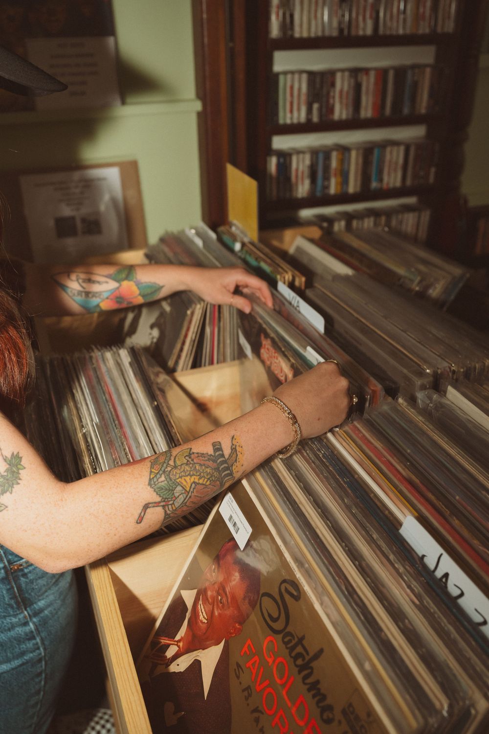 Tattooed hands browsing through a collection of vinyl records in a home library with wooden shelves.