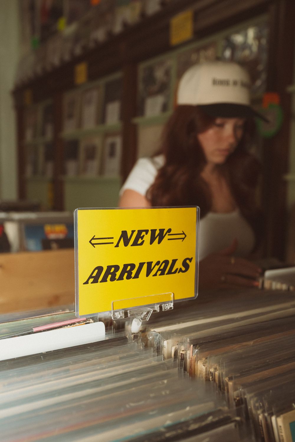 Yellow 'New Arrivals' sign displayed above vinyl record bins in a dimly lit record store.