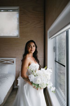 A bride in a classic white wedding gown holds a luxurious white bouquet while standing by a window.