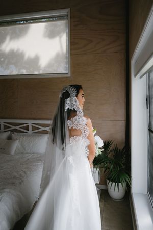 Elegant bride in lace wedding dress stands by window in natural light creating a romantic silhouette.
