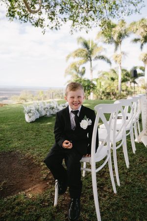 A ring bearer in a black suit sits playfully among white folding chairs at an outdoor wedding ceremony setup.