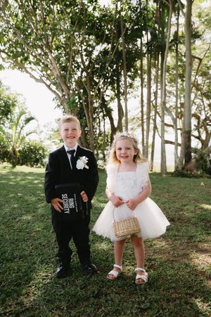 Two young children dressed in formal wedding attire stand together on a grassy lawn with trees in the background.