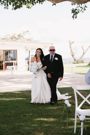 A wedding couple poses together outdoors on a sunny day with white chairs and greenery in the background.