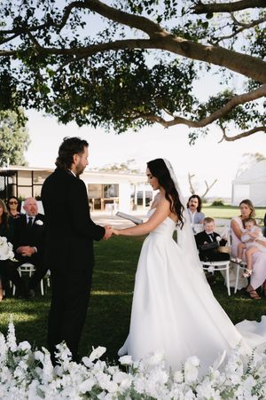 An outdoor wedding ceremony under a large tree with white floral arrangements decorating the aisle.