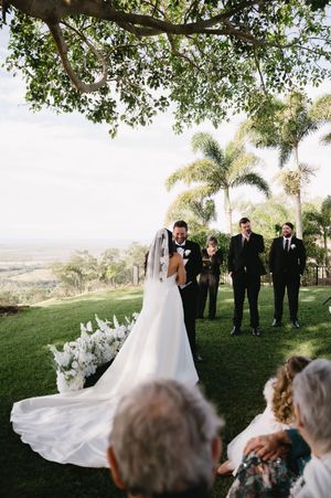 Wedding party poses under palm trees on a grassy hillside during an outdoor ceremony.