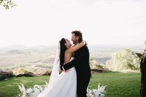 A couple embraces on a hilltop during their wedding ceremony with a scenic valley view in the background.