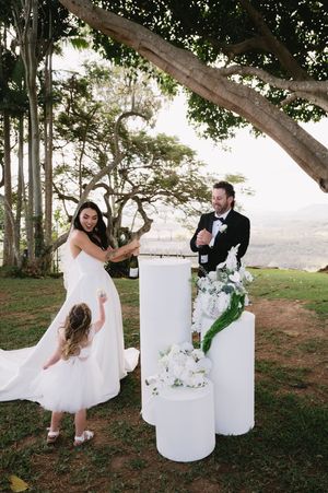 Wedding couple posing with elegant white pedestals and floral arrangements under a tree canopy.