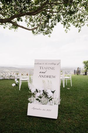 Elegant wedding welcome sign with white flowers stands on grassy hilltop under tree branches with scenic view.