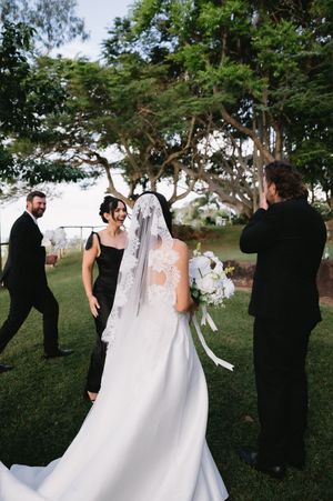 A wedding party gathered under trees with flowing white dress and formal attire visible in the outdoor setting.