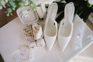 Elegant white pointed-toe bridal shoes displayed with jewelry and accessories on a light surface.