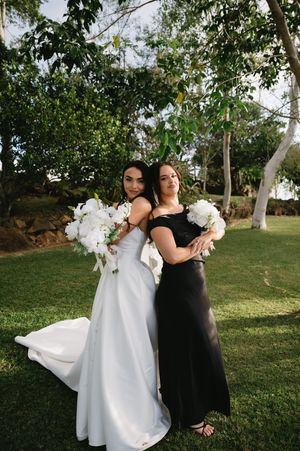A bride in white and woman in black stand together holding bouquets in an outdoor garden setting.