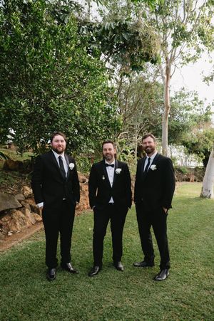 Groomsmen in black tuxedos pose together on grass beneath tall trees.