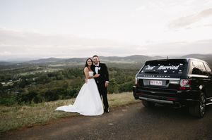 A wedding couple poses near their luxury vehicle with a scenic mountain backdrop.