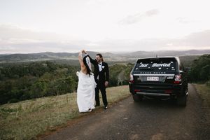 Newlyweds celebrate next to their black Range Rover on a scenic mountain road.