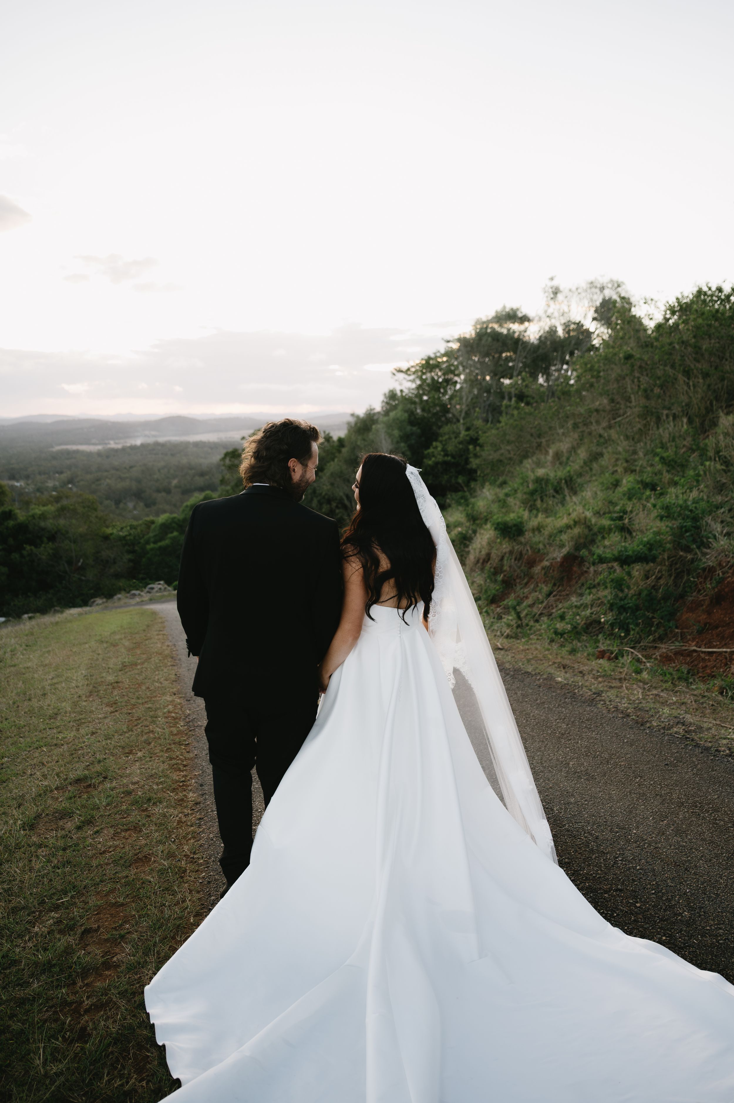 Sunset wedding photos on hilltop path with flowing white dress trailing behind.