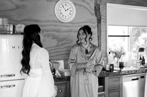 Two people in robes chat in a bright kitchen with a wall clock visible in the background.