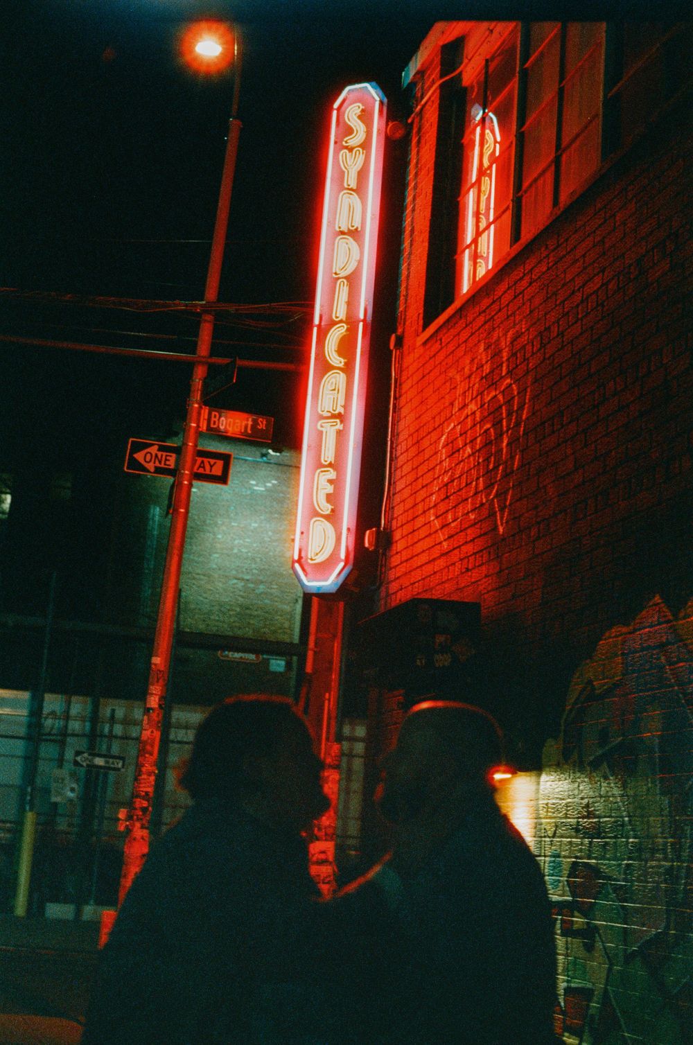 Two silhouetted figures stand beneath a glowing red neon Syndicated sign on a brick building at night.