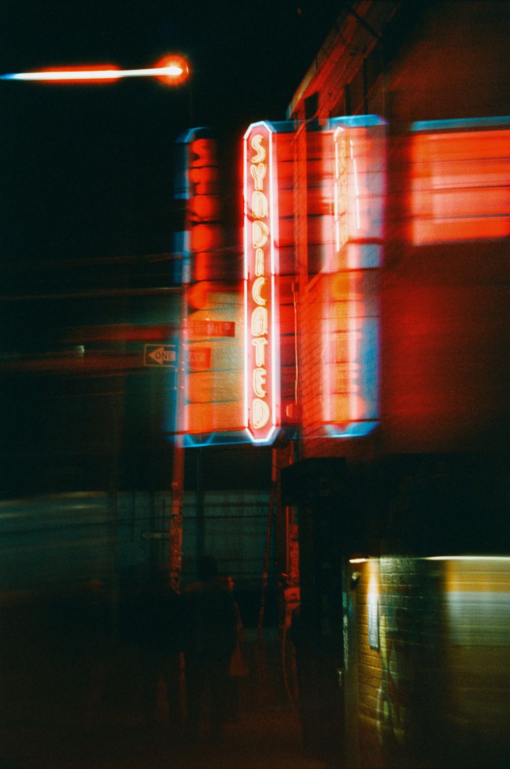 Blurry neon signs glow red on a dark city street at night, captured with motion blur effect.