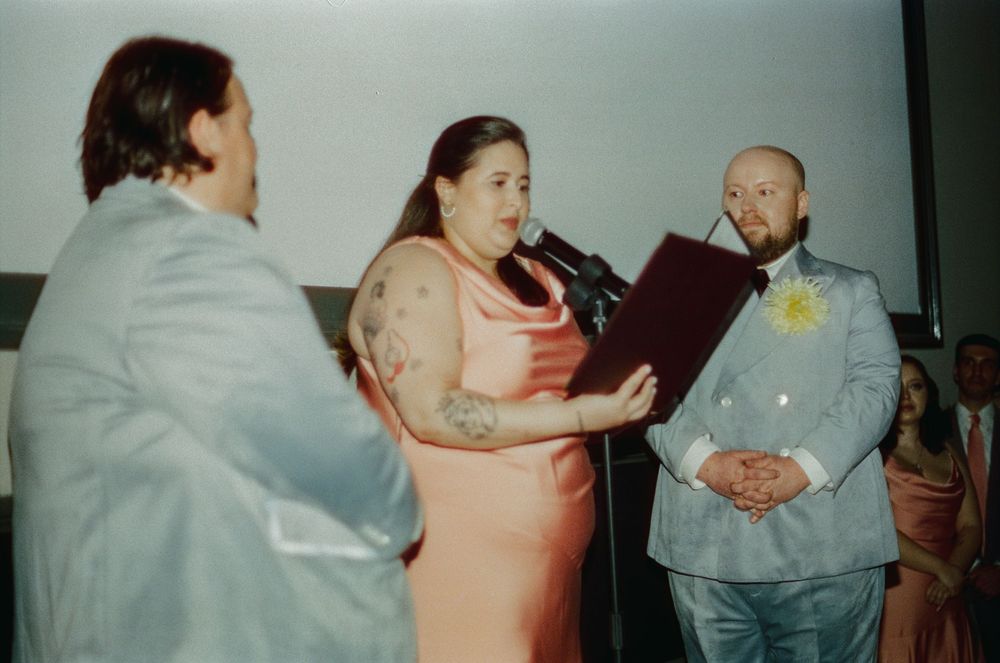 Wedding ceremony with tattooed officiant in pink dress reading vows, groom in light blue suit with yellow boutonniere.