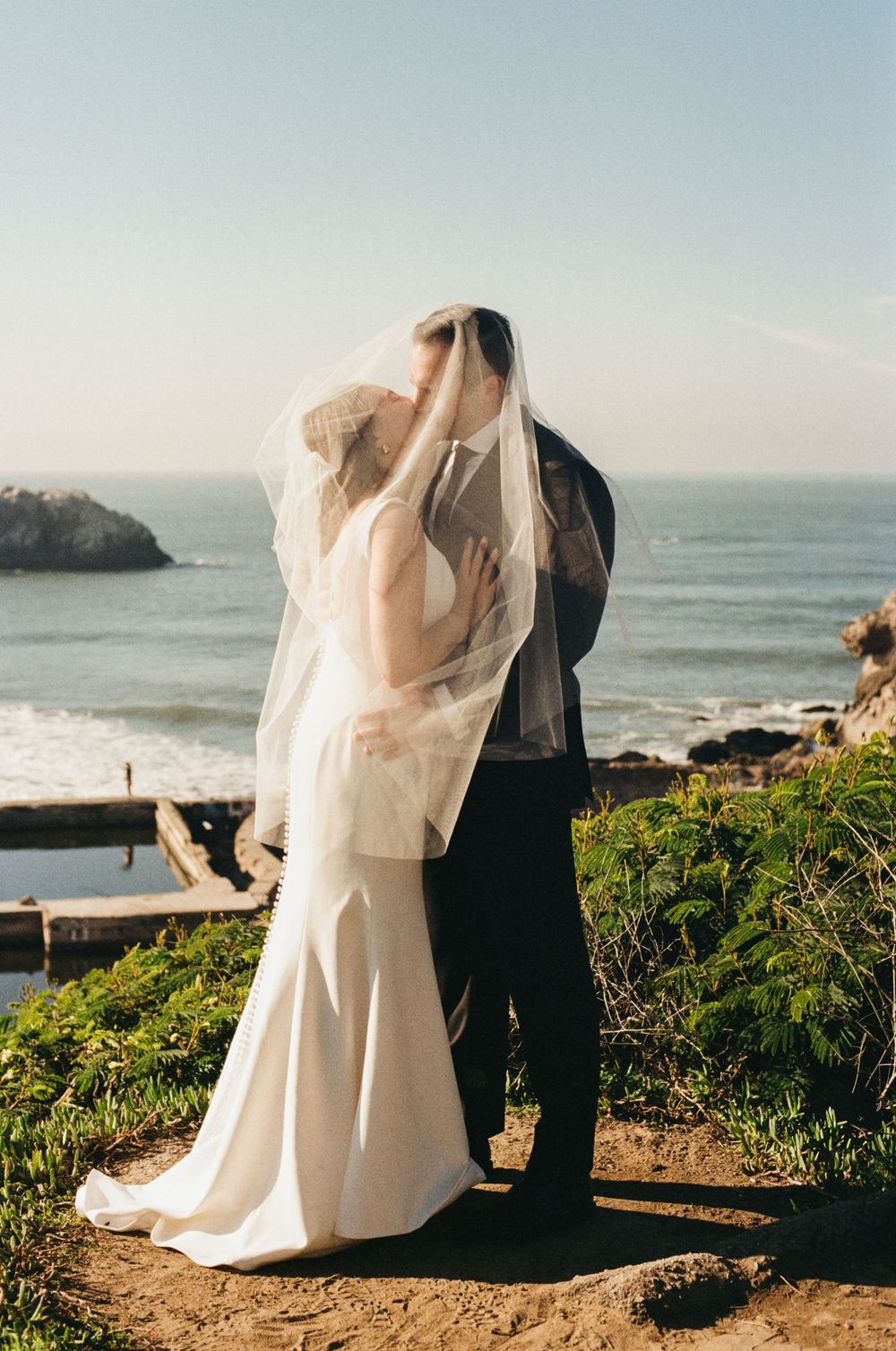 A romantic coastal wedding photoshoot featuring a couple against a scenic ocean backdrop with cliffs and greenery.