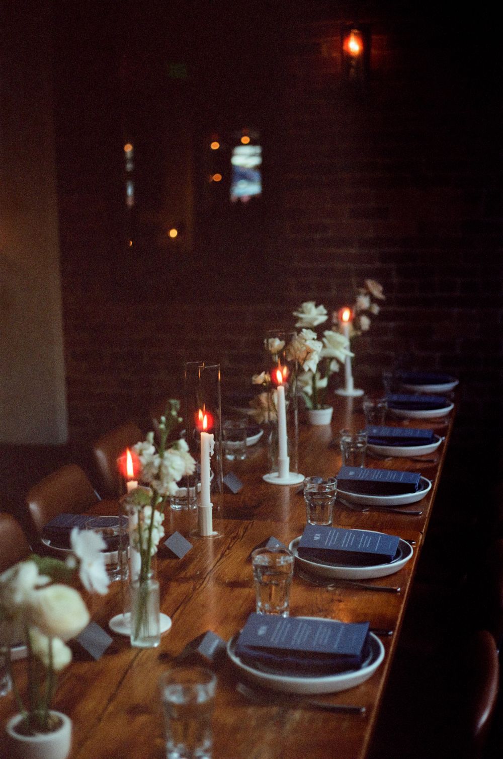 A rustic wooden dining table set with dark plates, candles and simple decor in moody lighting.