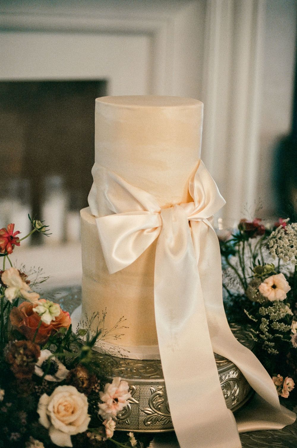 Elegant white wedding cake decorated with cream ribbon and surrounded by delicate floral arrangements on crystal stands.
