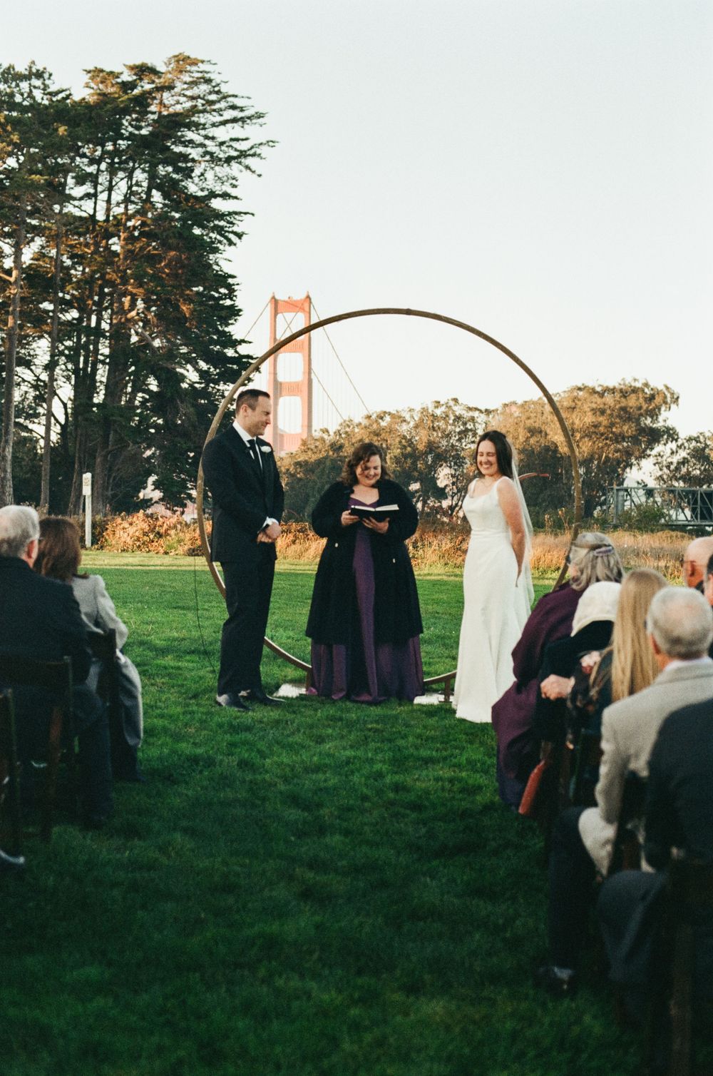 Outdoor wedding ceremony under an arch with the Golden Gate Bridge visible in the background.