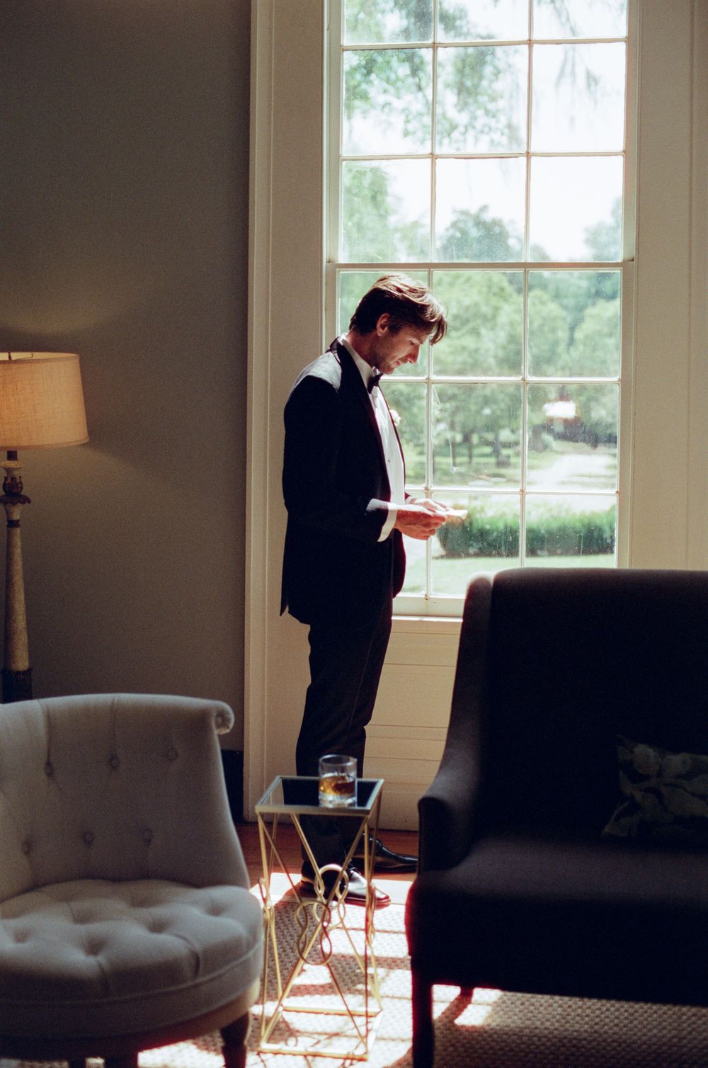 A figure in formal attire stands pensively by large windows in an elegant room with grey walls and modern furniture.