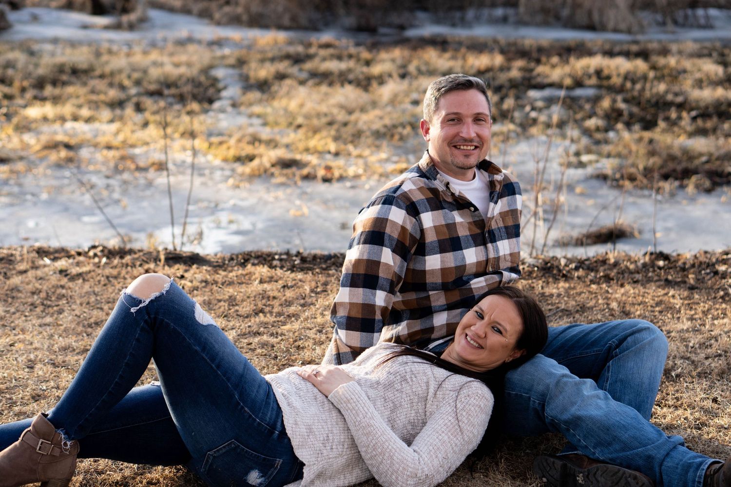 Smiling couple posing in a winter field, woman reclining against man, both wearing casual jeans and sweaters.