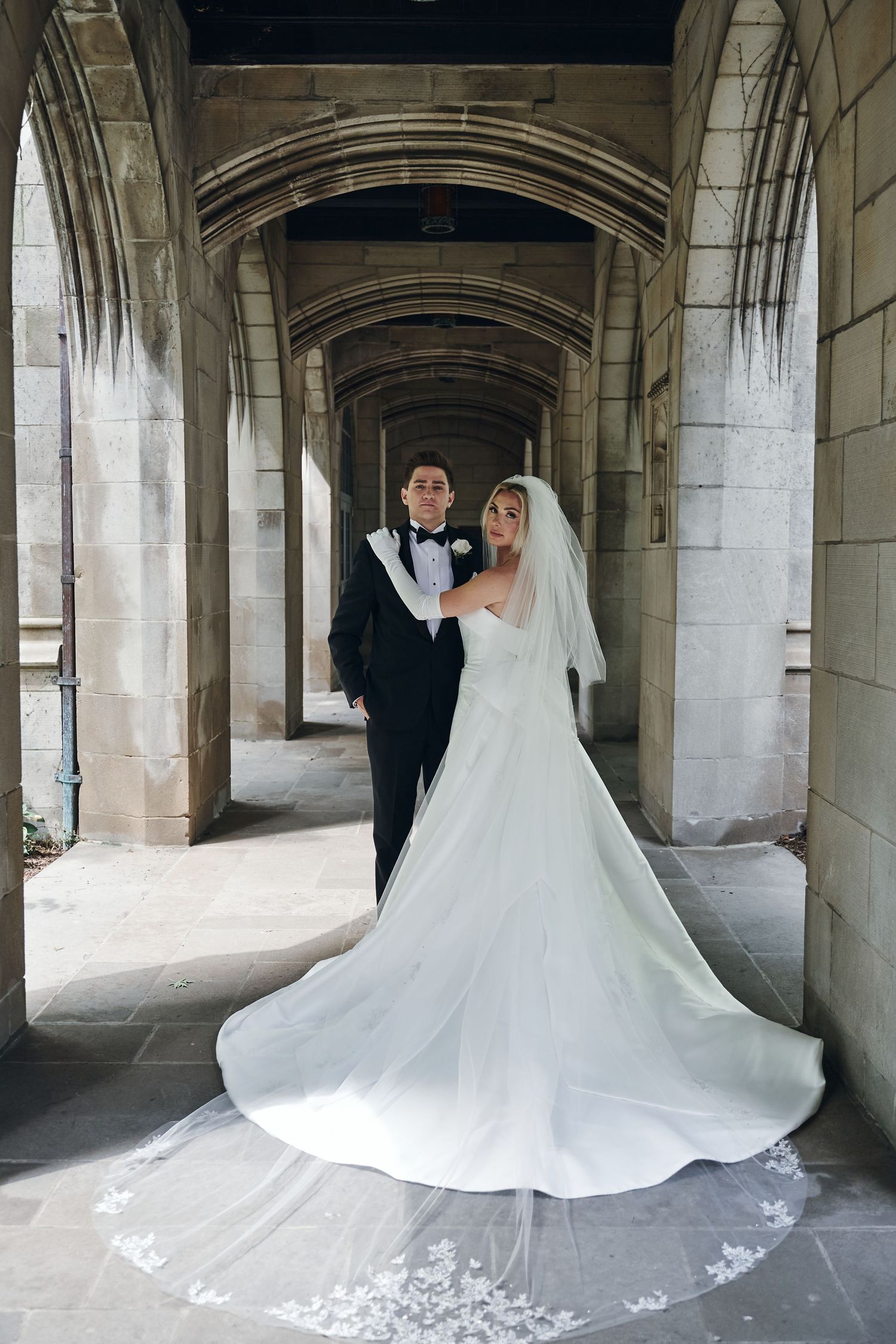 A wedding couple poses in a dramatic stone archway corridor with a flowing white bridal gown creating elegant movement.