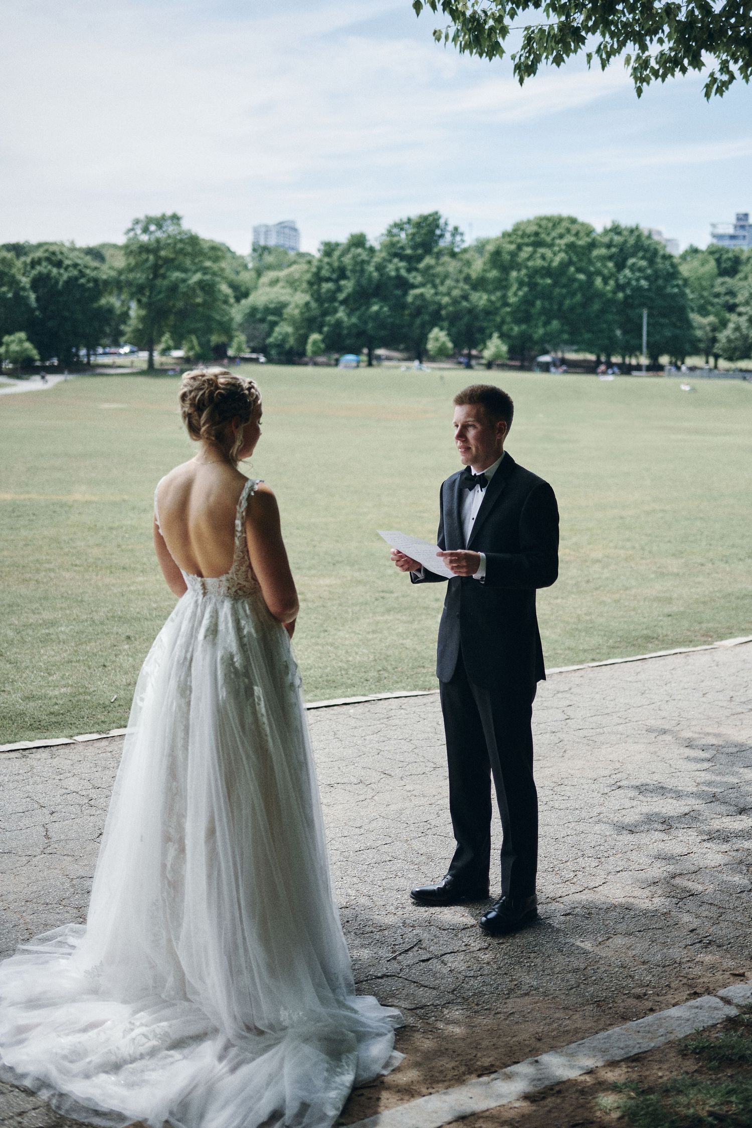 A couple in wedding attire stands in a park, with the bride in a flowing white dress and the groom in a dark suit.
