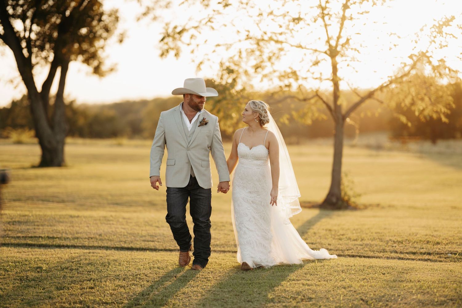 A couple walks hand in hand through a sun-drenched field during golden hour with trees in the background.