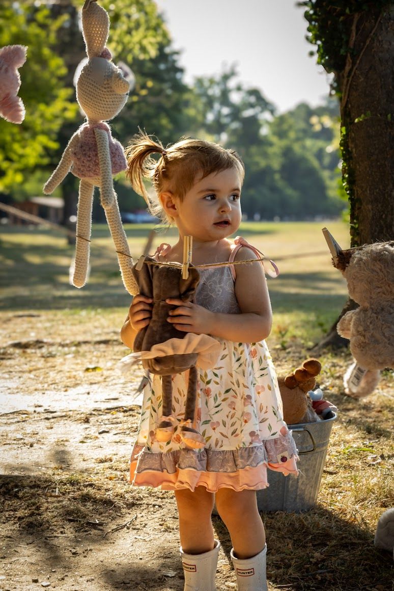 1 and a half year old girl in pigtails hanging stuffed animals out to dry wearing a dress with rain boots
