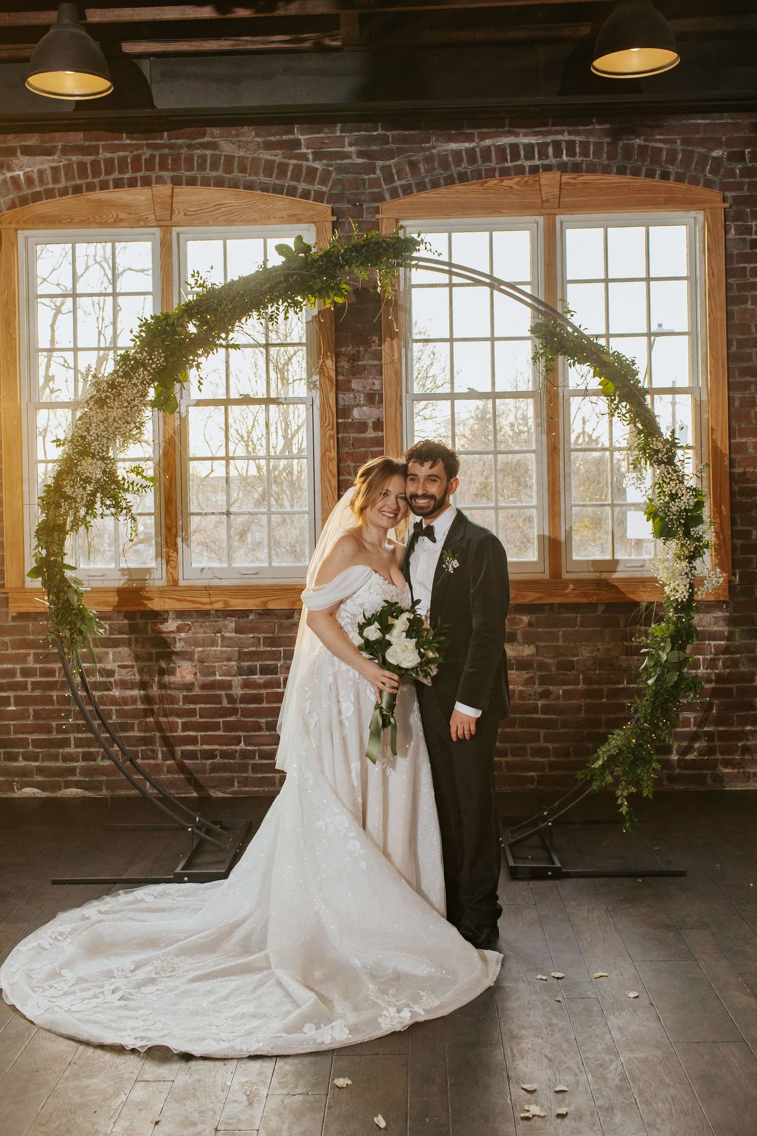 Romantic wedding couple embraces in rustic brick venue with circular floral arch and natural window light.