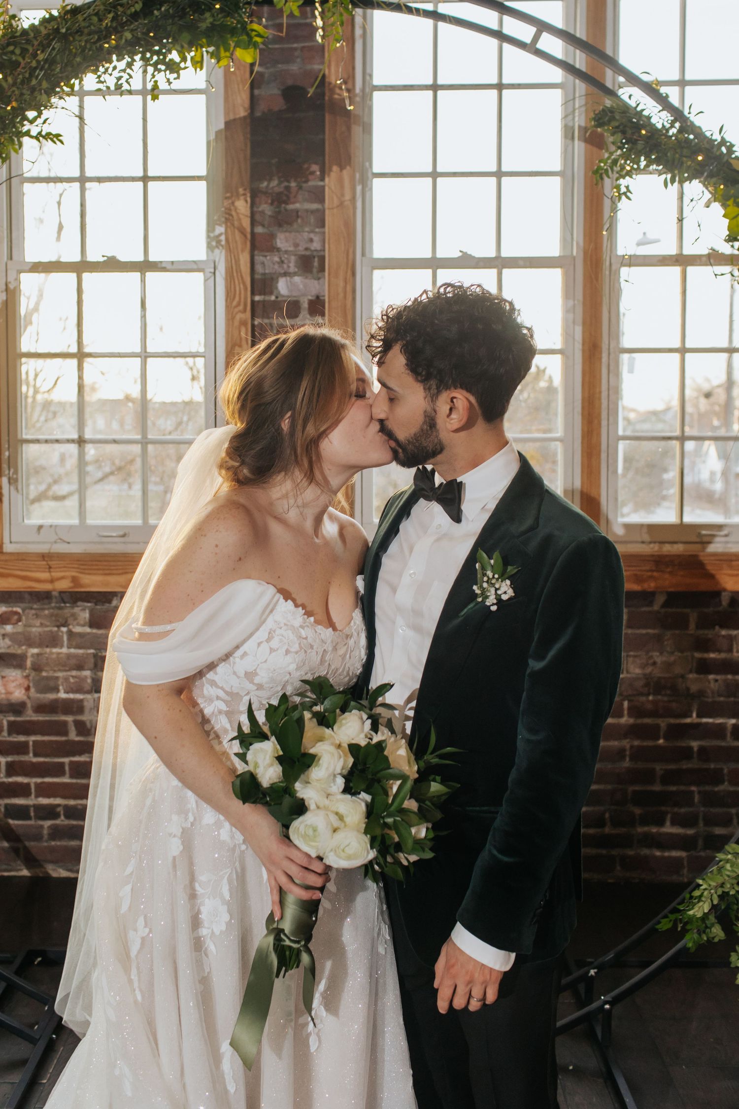 Romantic wedding couple shares a tender kiss in front of exposed brick wall and sunlit windows with greenery.