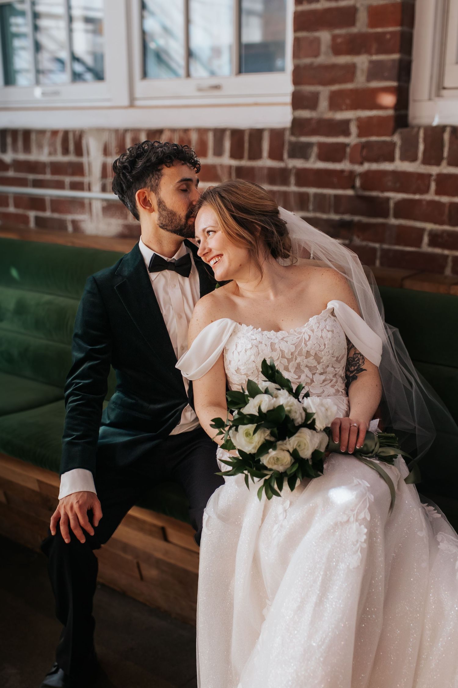 Romantic wedding couple shares intimate moment on vintage green velvet couch against exposed brick wall.