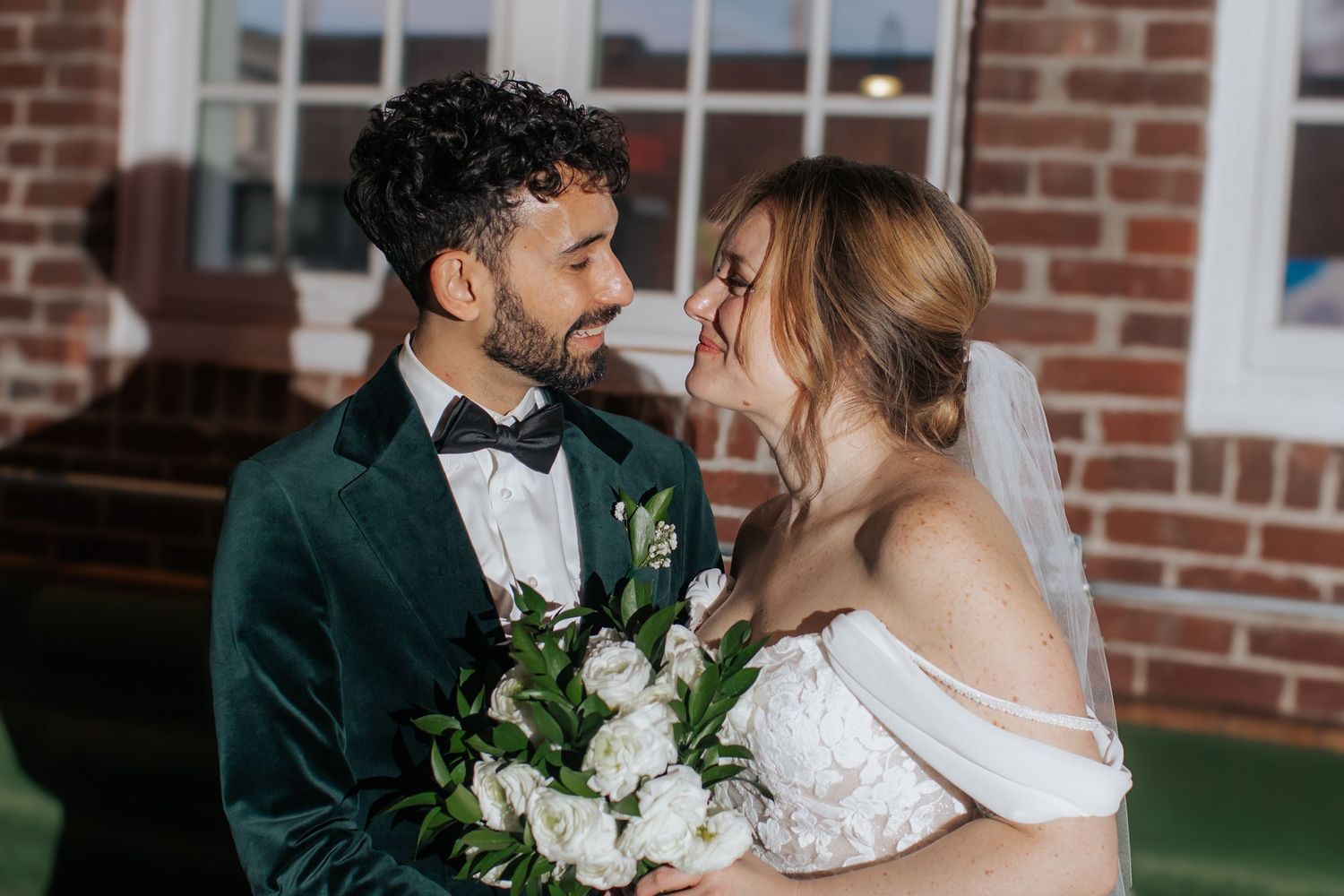 Romantic wedding couple embraces against rustic brick wall with white floral bouquet and elegant formal attire.
