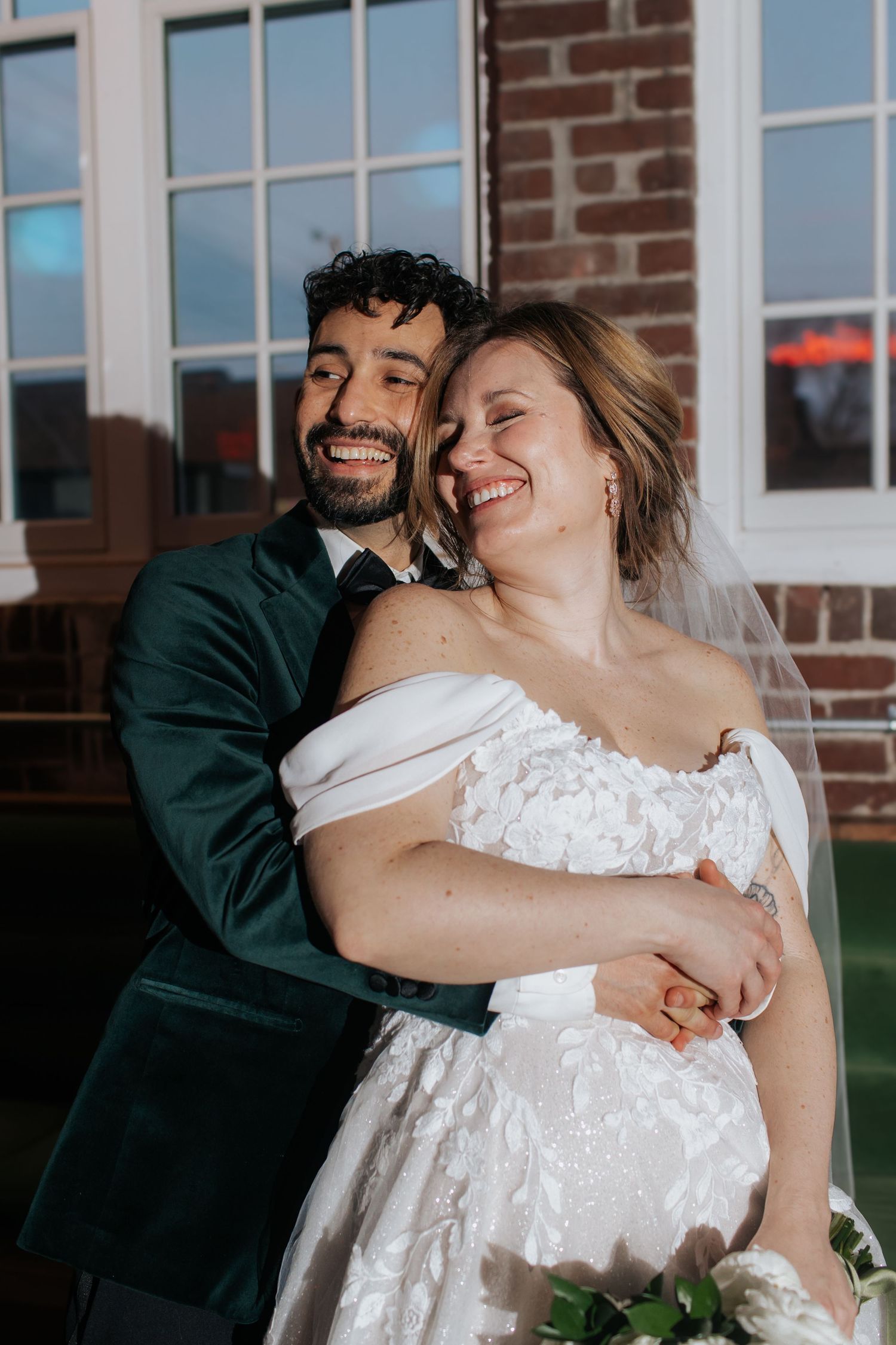 A joyful couple shares an intimate embrace outdoors against a brick building on their wedding day.