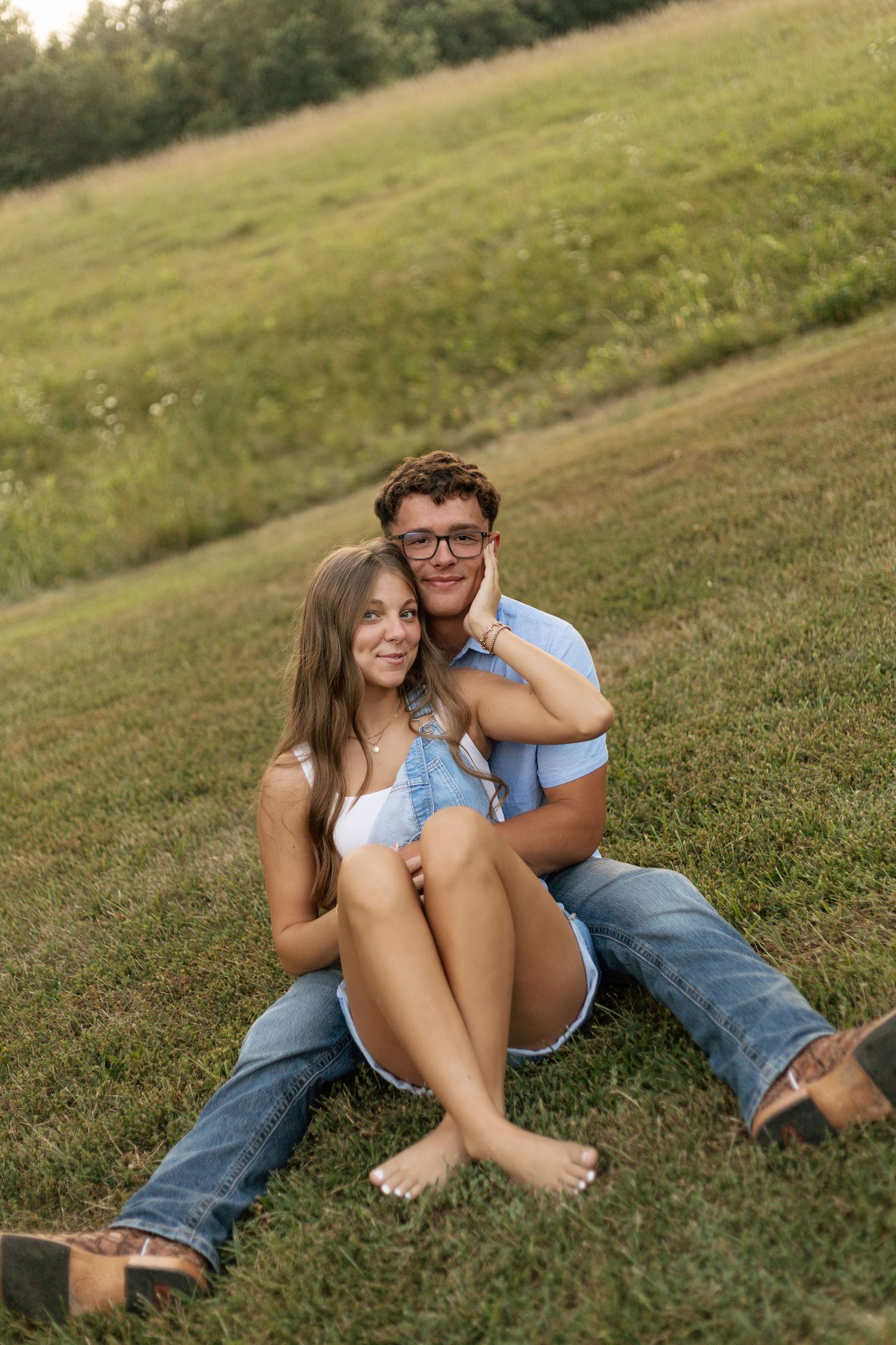 Couple embracing and smiling in golden hour sunlit field with trees.