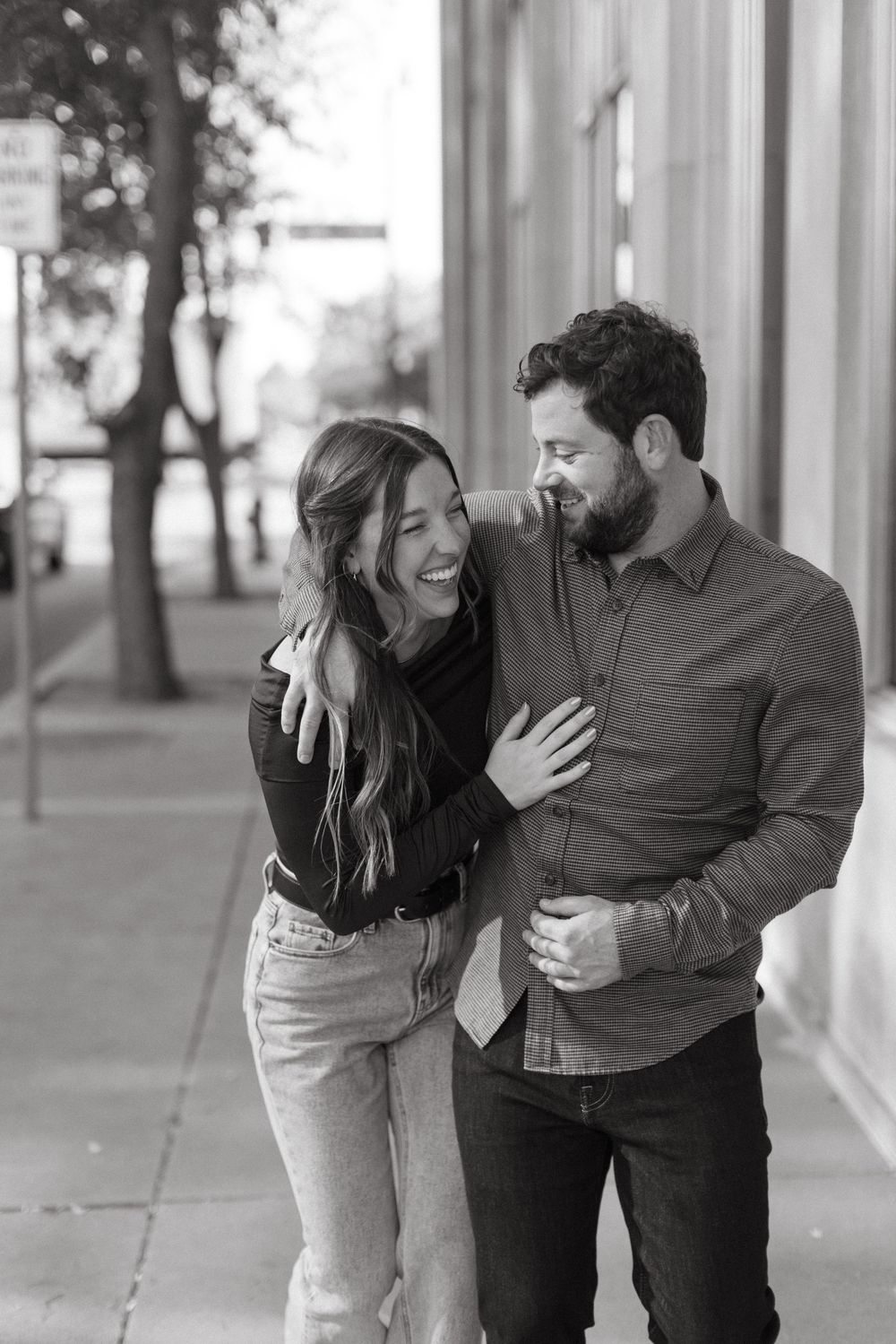 A couple laughs together while walking down a city sidewalk in black and white photography.