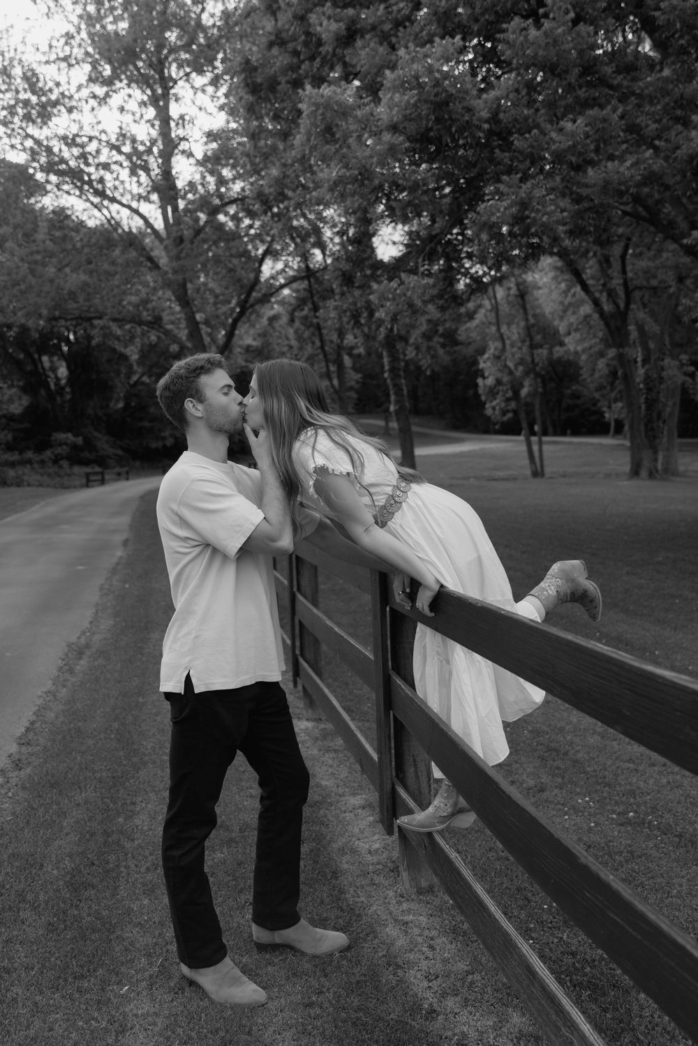 A romantic couple shares a tender moment leaning over a wooden fence in a park setting at dusk.