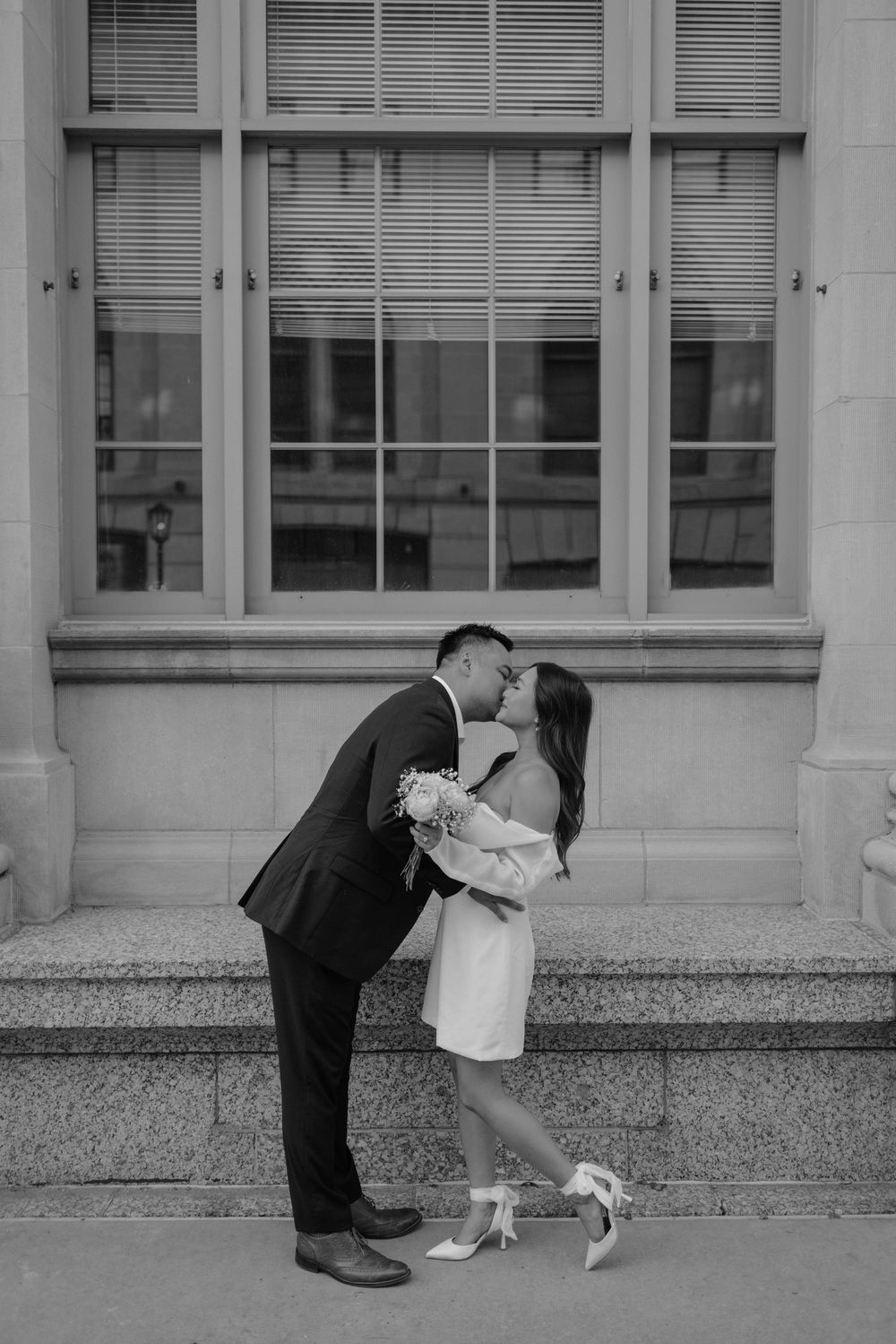 A romantic black and white photo of a couple sharing a kiss outside a building with large windows.