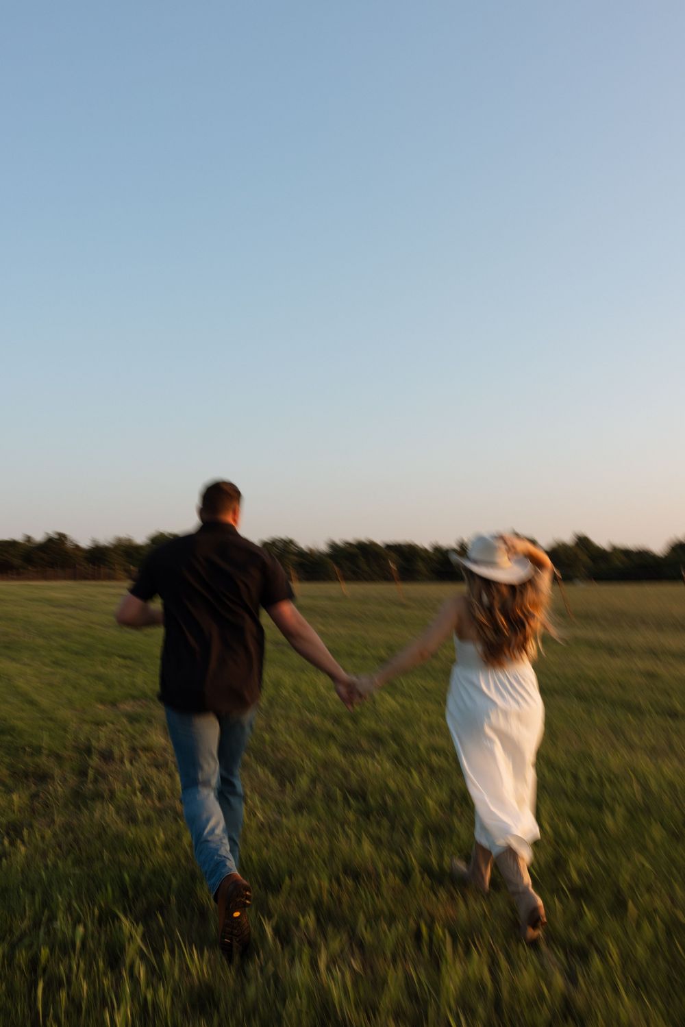 A couple runs playfully through a green field at sunset, holding hands and enjoying the outdoors together.