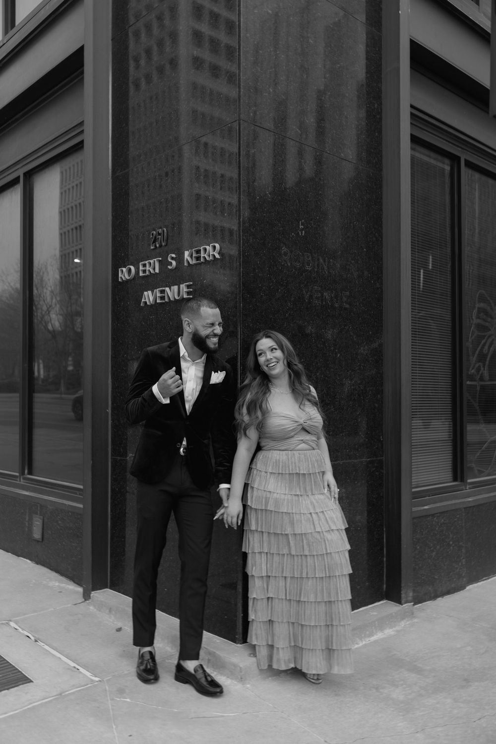A couple shares a playful moment outside a building entrance, with the woman wearing a tiered dress.