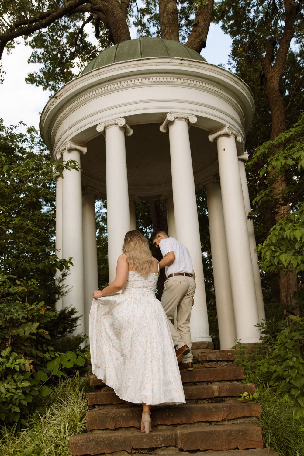 A couple ascends stone steps leading to a white classical gazebo surrounded by lush greenery.