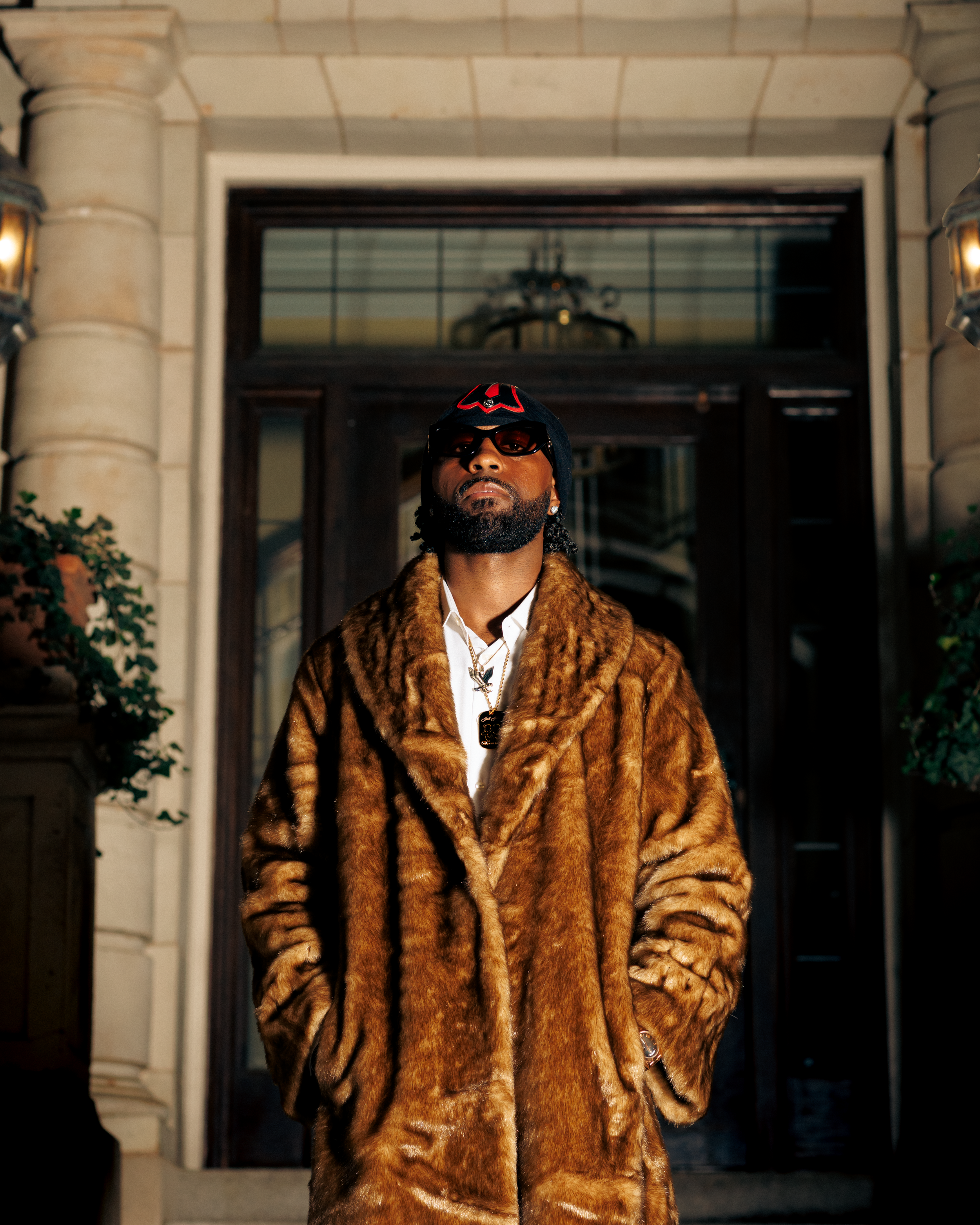 A person in a brown fur coat and baseball cap poses against an architectural backdrop with dramatic lighting.